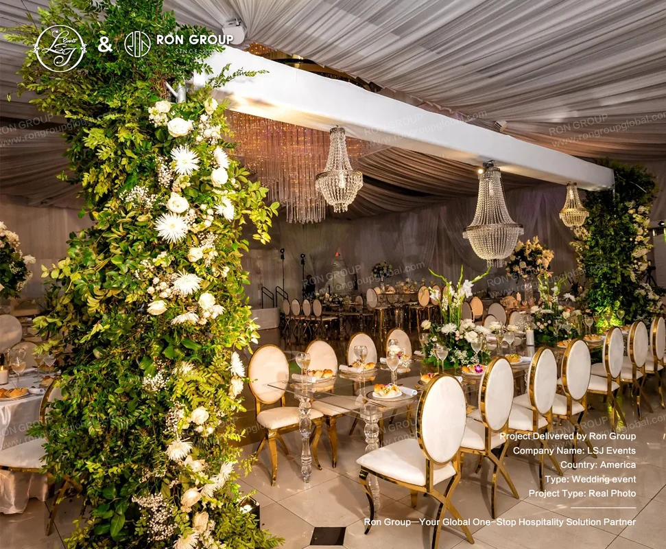 Luxurious wedding reception setup with glass tables, gold and cream chairs, greenery wall, and white floral arrangements