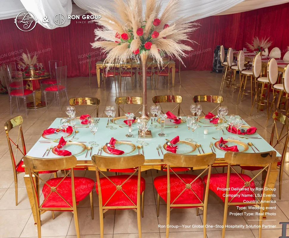 Wedding table setup with blue tabletop, gold-rimmed plates, red napkins, pampas grass, red roses, and gold chairs