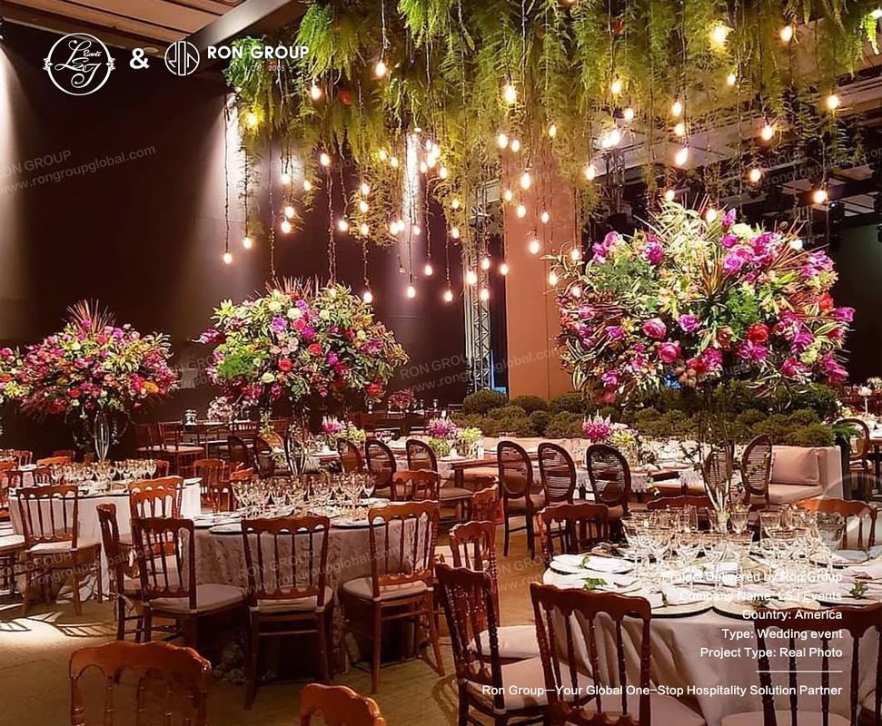Wedding reception round tables with white linens, wooden chairs, large floral centerpieces, greenery ceiling decor, and warm lights