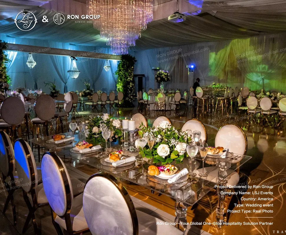 Glass reception tables with white floral centerpieces, candlelight, gold-trimmed chairs, chandelier lighting, and blue-green event lighting