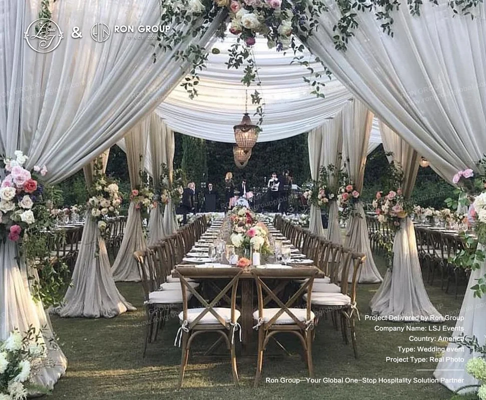 Outdoor wedding reception under a draped white tent with long wooden tables, floral arrangements, and rustic chairs