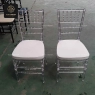 Two clear resin Chiavari chairs with white removable pads in a warehouse.