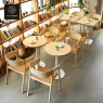 A cafe setting with multiple light wood chairs and round/square tables amidst bookshelves.
