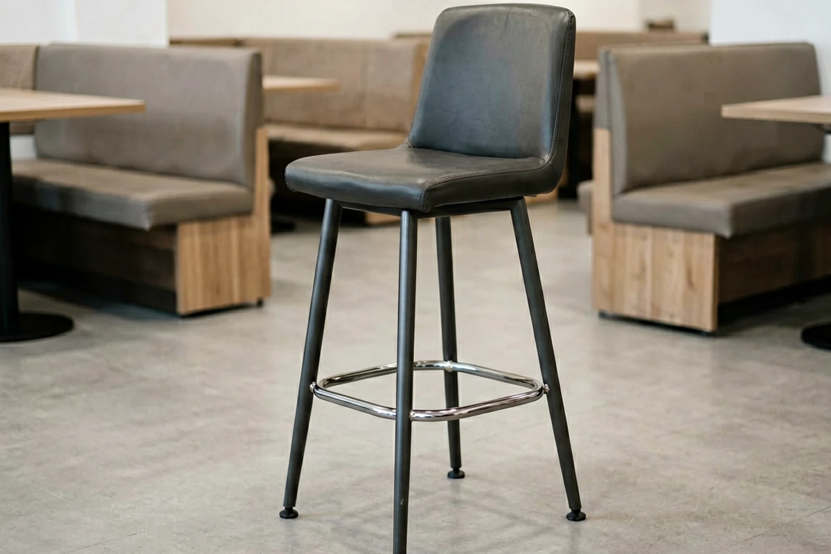 A modern black leather restaurant bar stool with metal legs and footrest standing in a dining area.